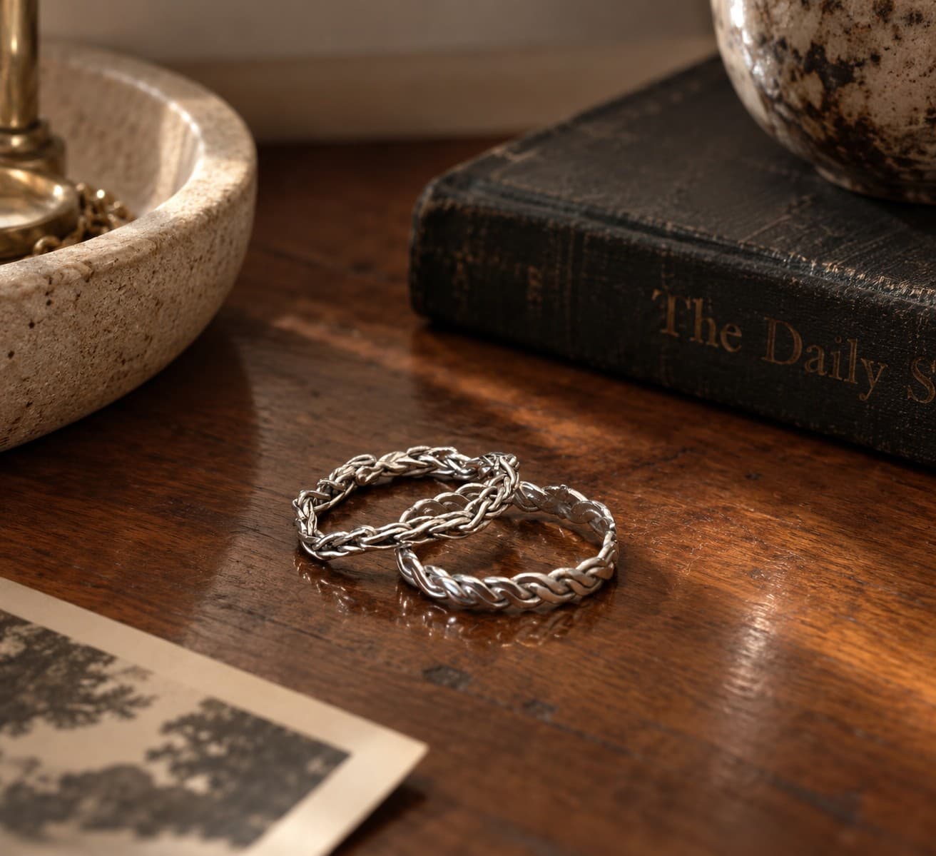 Two braided metal rings resting on a pale surface, catching warm daylight along the twist.